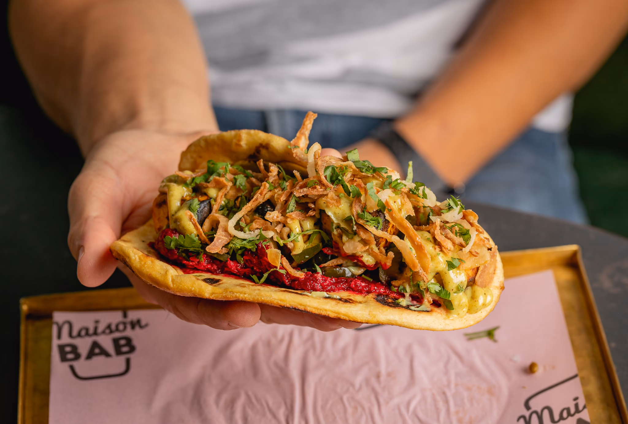 Hand holding pita bread stuffed with fries, greens, and drizzled white sauce.